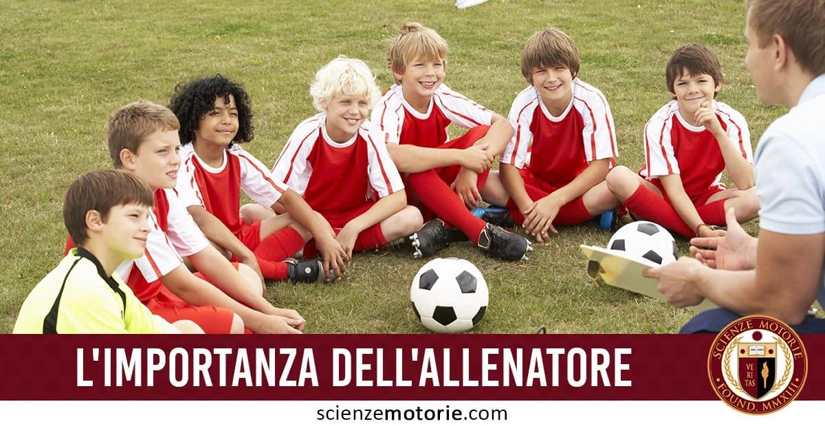 Bambini seduti in cerchio sull'erba, con maglie da calcio rosse e bianche, ascoltano un allenatore che tiene un blocco note, sul campo da calcio. l'immagine è accompagnata dalla scritta "L’importanza dell’allenatore" e dal logo di ScienzeMotorie.com