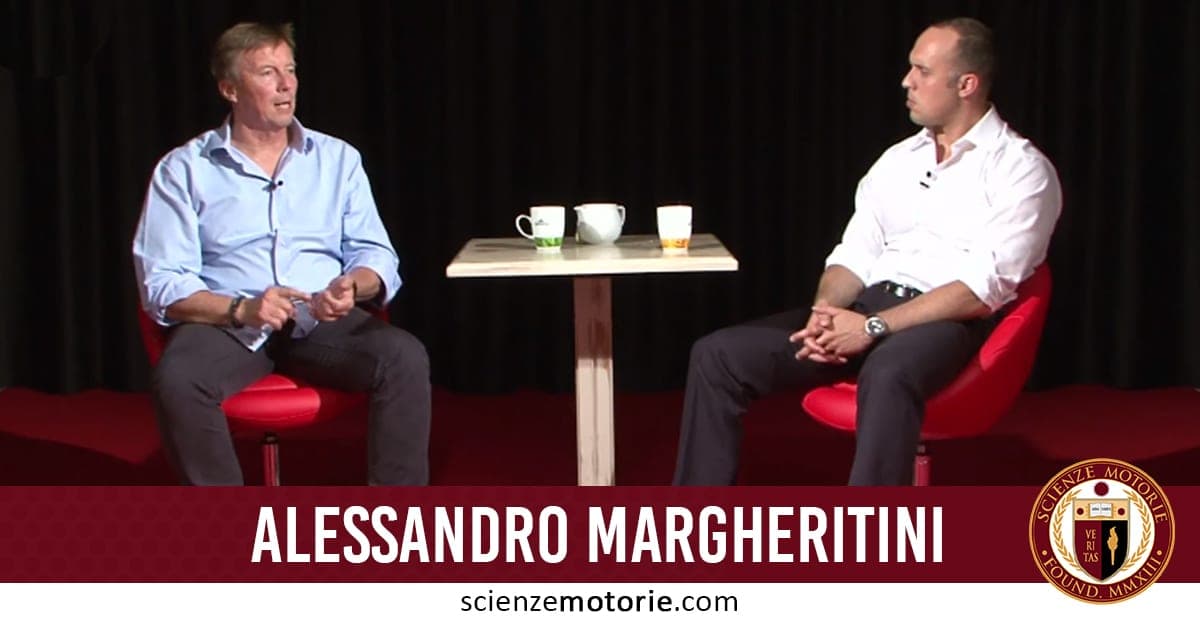 Due uomini seduti su sedie rosse su un palco di fronte a un tavolo con tre tazze. A sinistra Alessandro Margheritini con camicia azzurra e pantaloni scuri, a destra Giacomo Catalani con camicia bianca. Sullo sfondo un ambiente scuro, con scritta "Alessandro Margheritini" e logo scienzemotorie.com in basso.