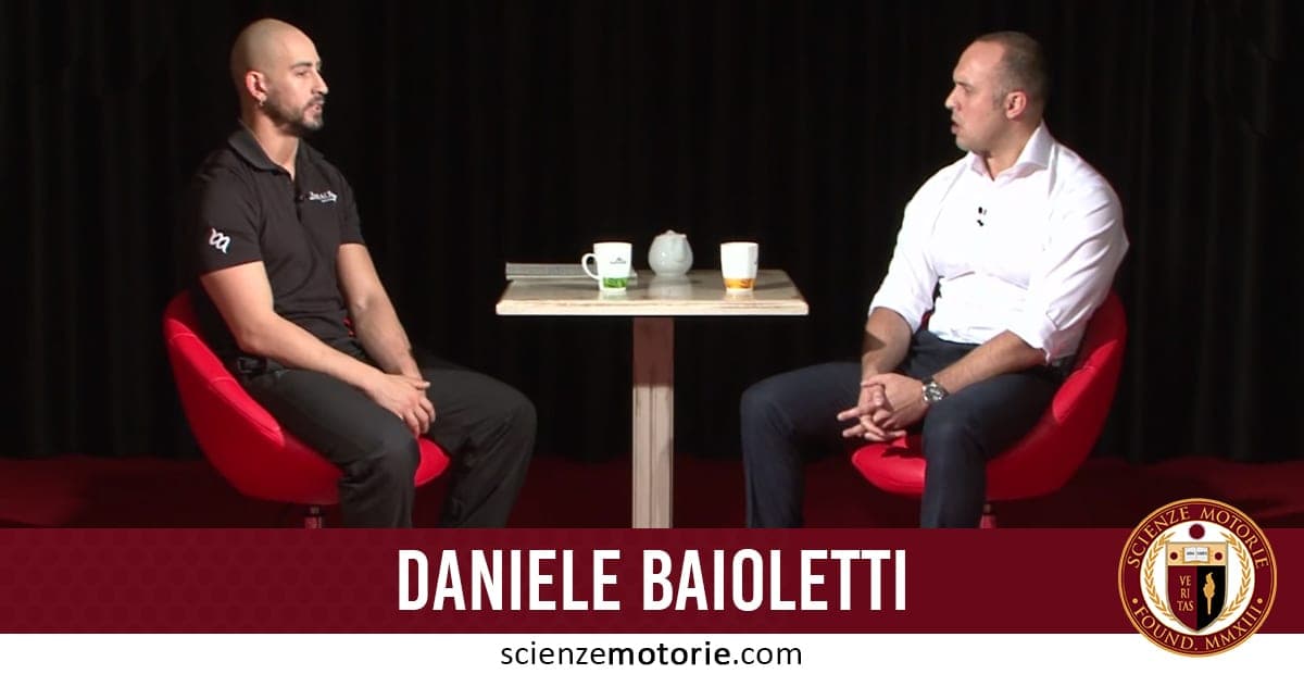 Daniele Baioletti e Giacomo Catalani seduti a un tavolo durante un'intervista per Scienze Motorie. Entrambi in conversazione, con tazze sul tavolo e tenda nera sullo sfondo.