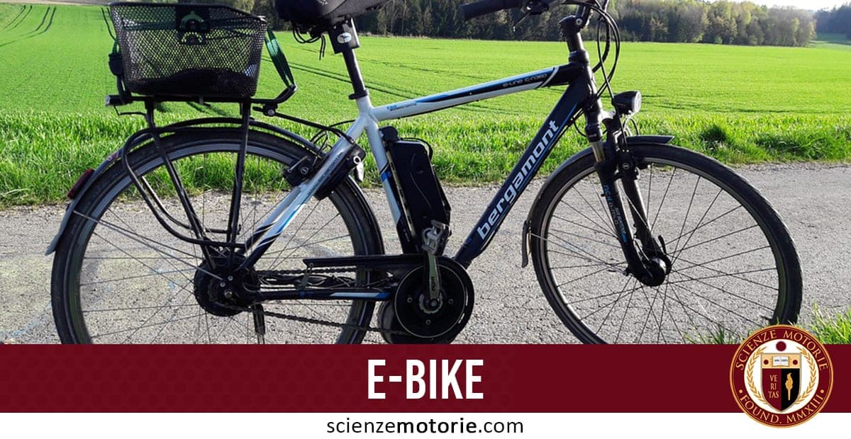 Bicicletta elettrica nera e blu con cestino posteriore parcheggiata su una strada di campagna; in basso scritta "E-BIKE" e logo di ScienzeMotorie.com.