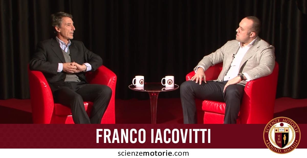 Franco Iacovitti e Giacomo Catalani seduti su poltrone rosse durante un’intervista con tavolino e tazze bianche, sfondo con tenda nera.