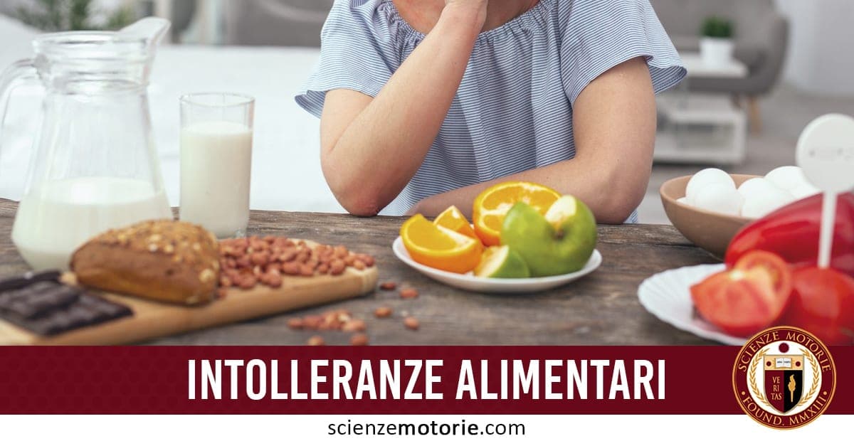 L'immagine mostra una persona seduta a un tavolo con vari alimenti: c'è un bicchiere e una caraffa di latte, un piatto con uova, un altro piatto con agrumi come arance e lime, e una tavoletta di cioccolato accanto a del pane e noci. Sul fondo c'è la scritta "INTOLLERANZE ALIMENTARI" e il logo "scienzemotorie.com".