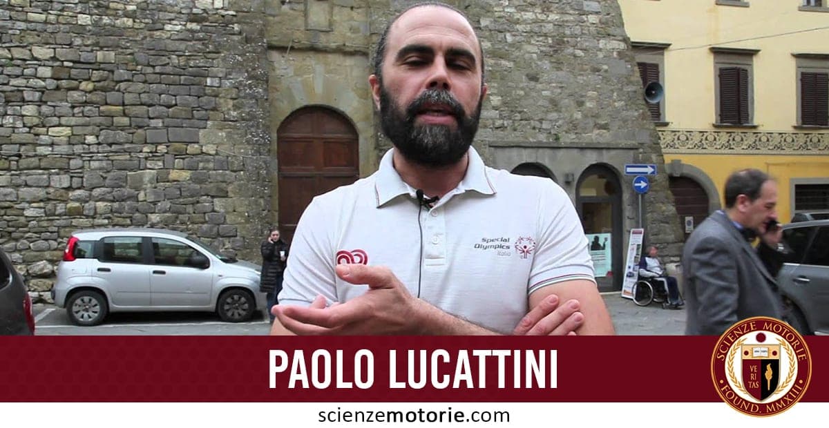 Uomo con barba in polo bianca davanti a un edificio di pietra con persone e una macchina parcheggiata, Paolo Lucattini.