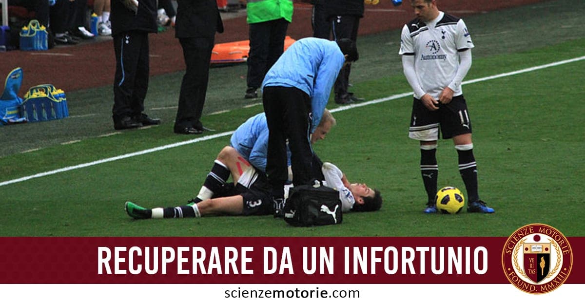 L’immagine mostra un calciatore con la maglia numero 3 a terra, infortunato, assistito da una persona in maglia azzurra con una borsa medica accanto. Un altro giocatore con la maglia bianca numero 10 osserva la scena. Sul prato si vede un pallone giallo e nero. In basso, una banda rossa con la scritta “RECUPERARE DA UN INFORTUNIO” e il log del sito scienzemotorie.com.
