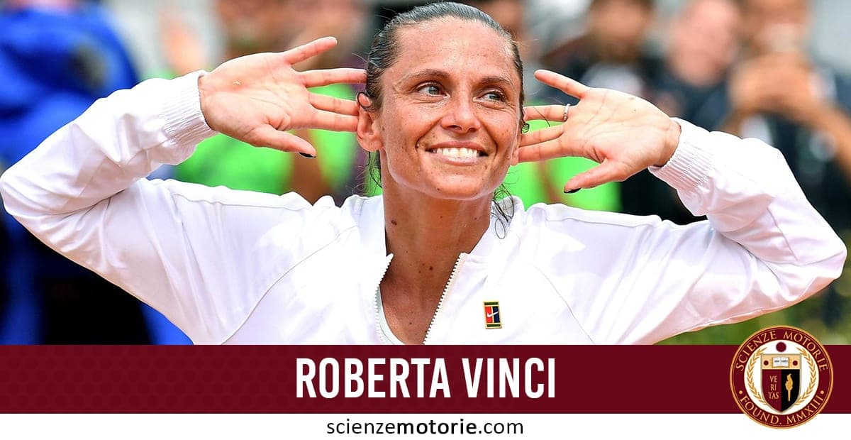 Roberta Vinci in primo piano con giacca sportiva bianca, sorridente con mani alzate in gesto di celebrazione. Logo ITF visibile sulla spalla destra della giacca. Sfondo sfocato di un campo da tennis.