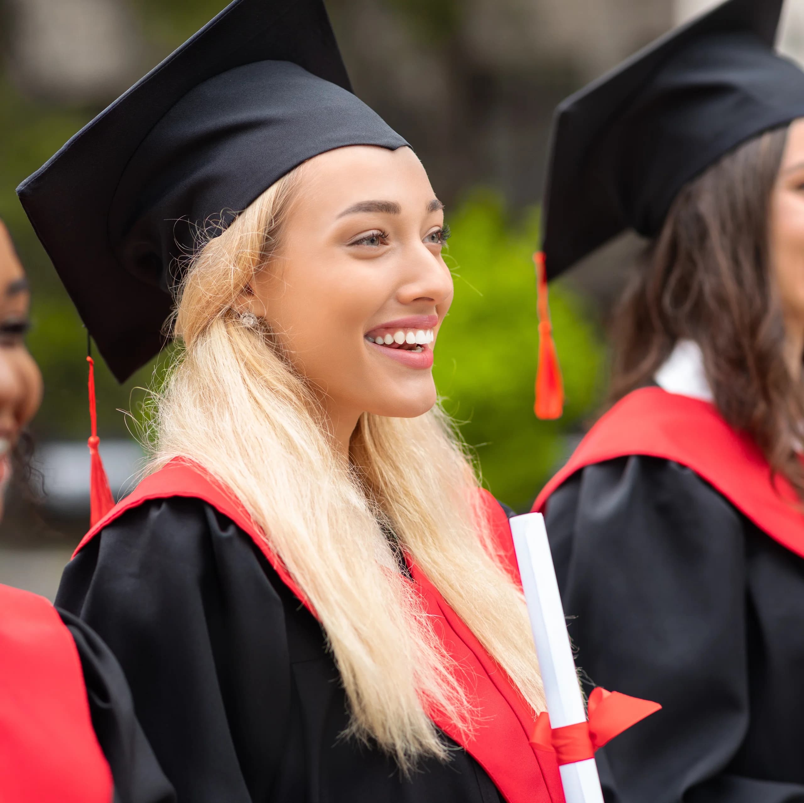 Studente sorridente con abito accademico nero, stola rossa e tocco che tiene un diploma avvolto con nastro rosso durante una cerimonia di laurea all’aperto.
